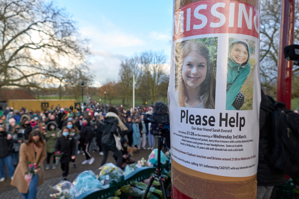 A missing poster featuring a photo of Sarah Everard appears on a post, with people laying flowers at a vigil in the background