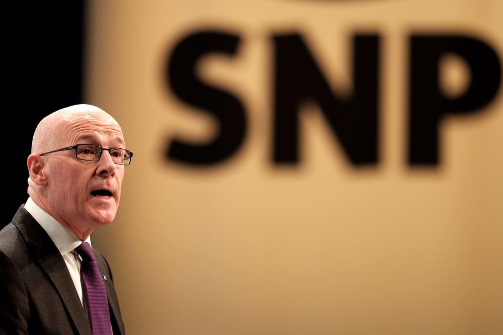 John Swinney stands in front of a yellow banner reading SNP