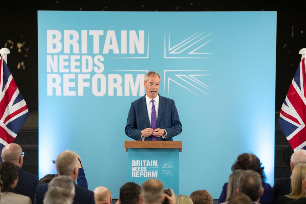 Nigel Farage stands at a lectern in front of a background reading Britain Needs Reform.