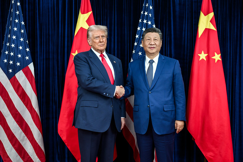 Donald Trump and Xi Jinping shake hands in front of Chinese and American flags.